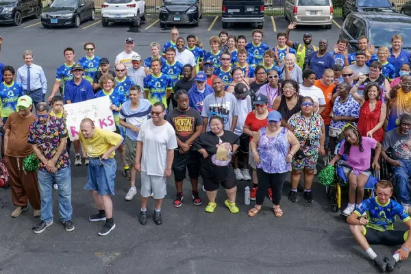 Large diverse group of over 100 people wearing blue 'Journey of Hope' shirts gathered in a parking lot, smiling and waving at the camera.