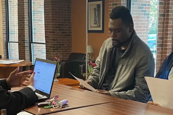 A woman with long black hair is on the left, seated at a wooden table, gesturing towards two men sitting across from her. She has a laptop open and a beige handbag placed on the back of the chair. The setting is a cozy indoor area with brick walls, large windows, and some wall art. The men are holding papers and engaging attentively.