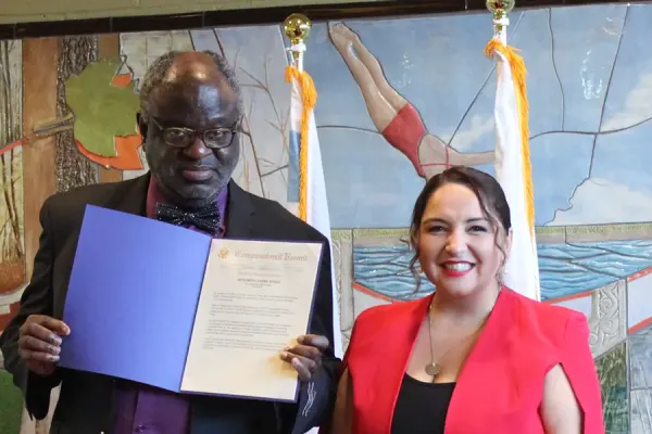 Two people standing with a U.S. House of Representatives podium and mural in the background. The man holds an open document. A vase of flowers is on a table between them.
