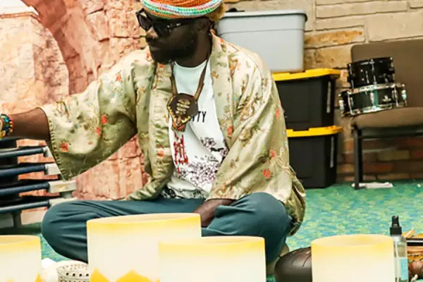 Person wearing a straw hat and patterned shirt demonstrating hand drums at a community healing event, with several yellow and white frame drums arranged on a teal and green carpeted floor.