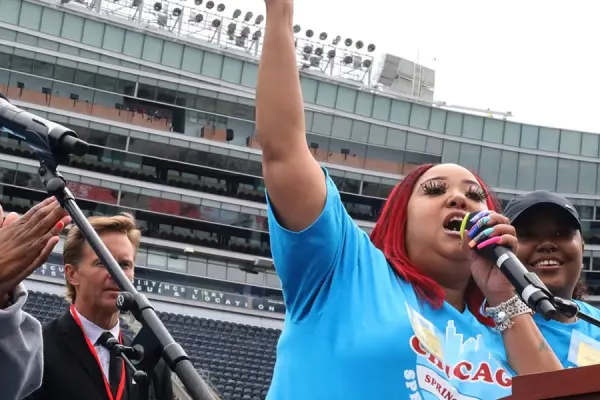 A person speaking energetically at a podium in a sports stadium with others nearby.