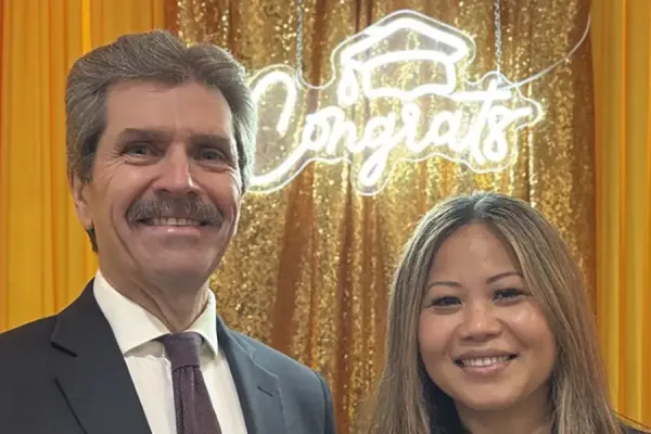 Two people smiling together at a celebratory event, posed in front of a gold sequin curtain and neon 'Congrats' sign, with colorful balloons nearby.
