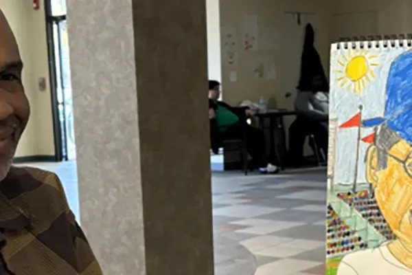 A man in a brown plaid shirt smiles while holding a colorful drawing of a person in a stadium, with colored pencils on the table.