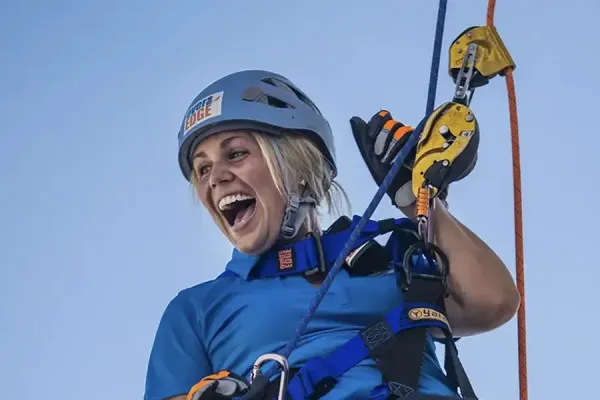 Person in blue safety gear and helmet smiling while rappelling outdoors, holding yellow and orange climbing carabiners attached to secured rope lines.