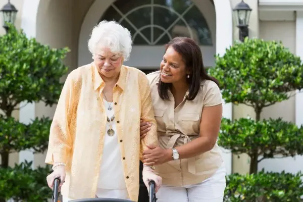 Older woman using a walker walks arm-in-arm with a smiling caregiver outside an elegant residential home