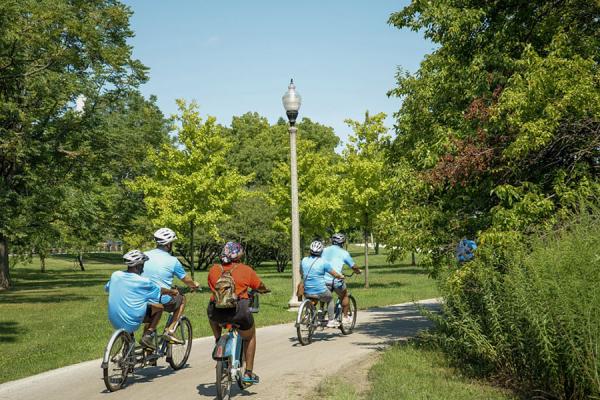 A group of cyclists bikes along a lakefront trail surrounded by green foliage and nature