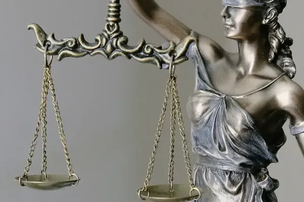 Metallic statue of Lady Justice in profile, holding balanced scales of justice in both hands, wearing a crown and classical robes against a neutral gray background.