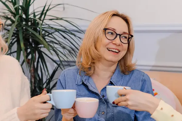 Three women smiling and toasting with coffee cups in an indoor office setting, conveying a moment of workplace camaraderie and celebration.