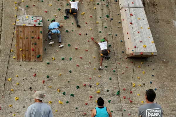 Six people at an indoor rock climbing wall; three climb the light brown wall with colorful holds while three others watch from the foreground.