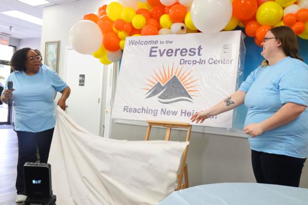 Two Envision Unlimited staff members reveal signage for the Everest drop-in center in a celebratory fashion, with balloons in the background