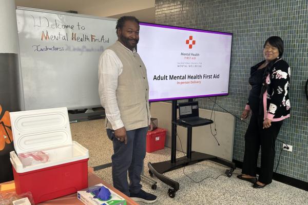 Two instructors stand in front of display materials regarding Mental Health First Aid training
