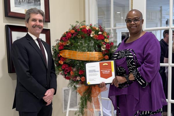 Two people celebrate a program graduation; a person in a suit and tie stands beside a floral arrangement on the left while a person in a purple dress with black and white accents holds a certificate on the right.