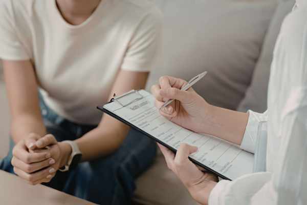 A person writes on a clipboard in the foreground while another sits on a couch leaning forward in the background.