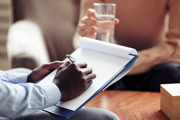 Two people meet across a table; one takes notes on a clipboard, the other holds a glass of water, with a tissue box between them.