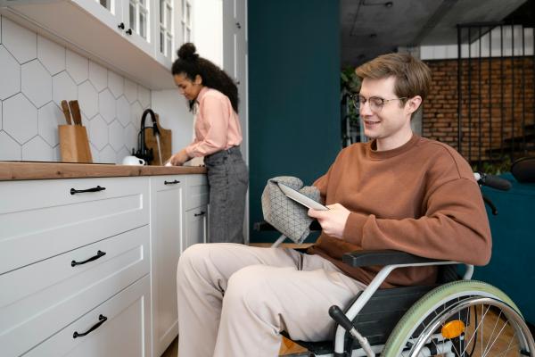 An individual is seen in the foreground in a wheelchair cleaning a dish, another individual assists in the background