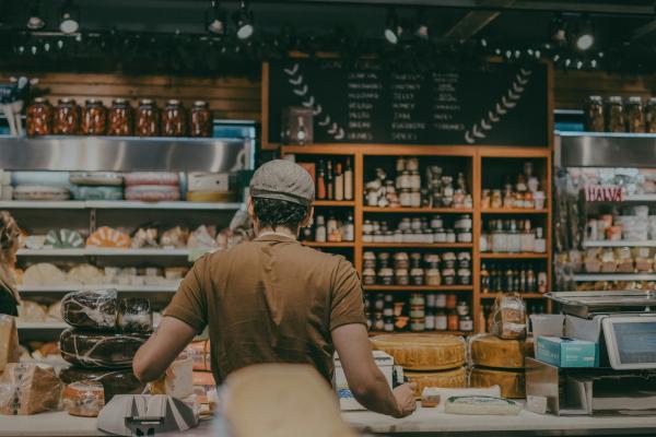 A person in a brown shirt and hat stands at a counter with shelves of food, jars, and cheese in the background.