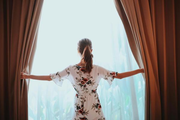A person in a white floral dress and ponytail opens light orange curtains at a bright, sunny window.