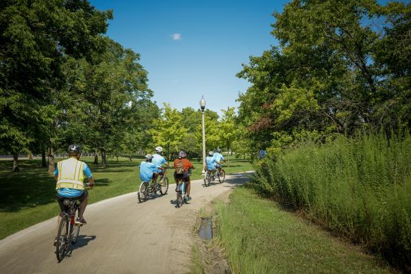 A group of individuals on different adaptive bicycles are pictured biking alongside a path with bright green foliage