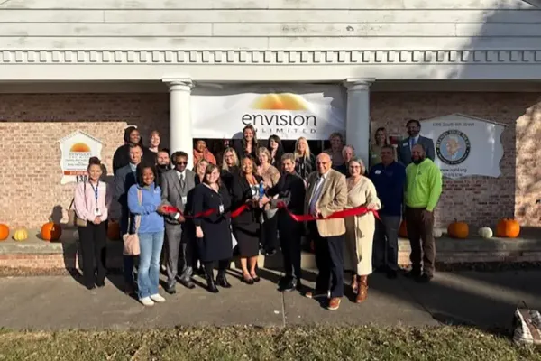 Group of community members and professionals cutting a red ribbon at the entrance of the Envision Unlimited mental health center building during its opening ceremony.