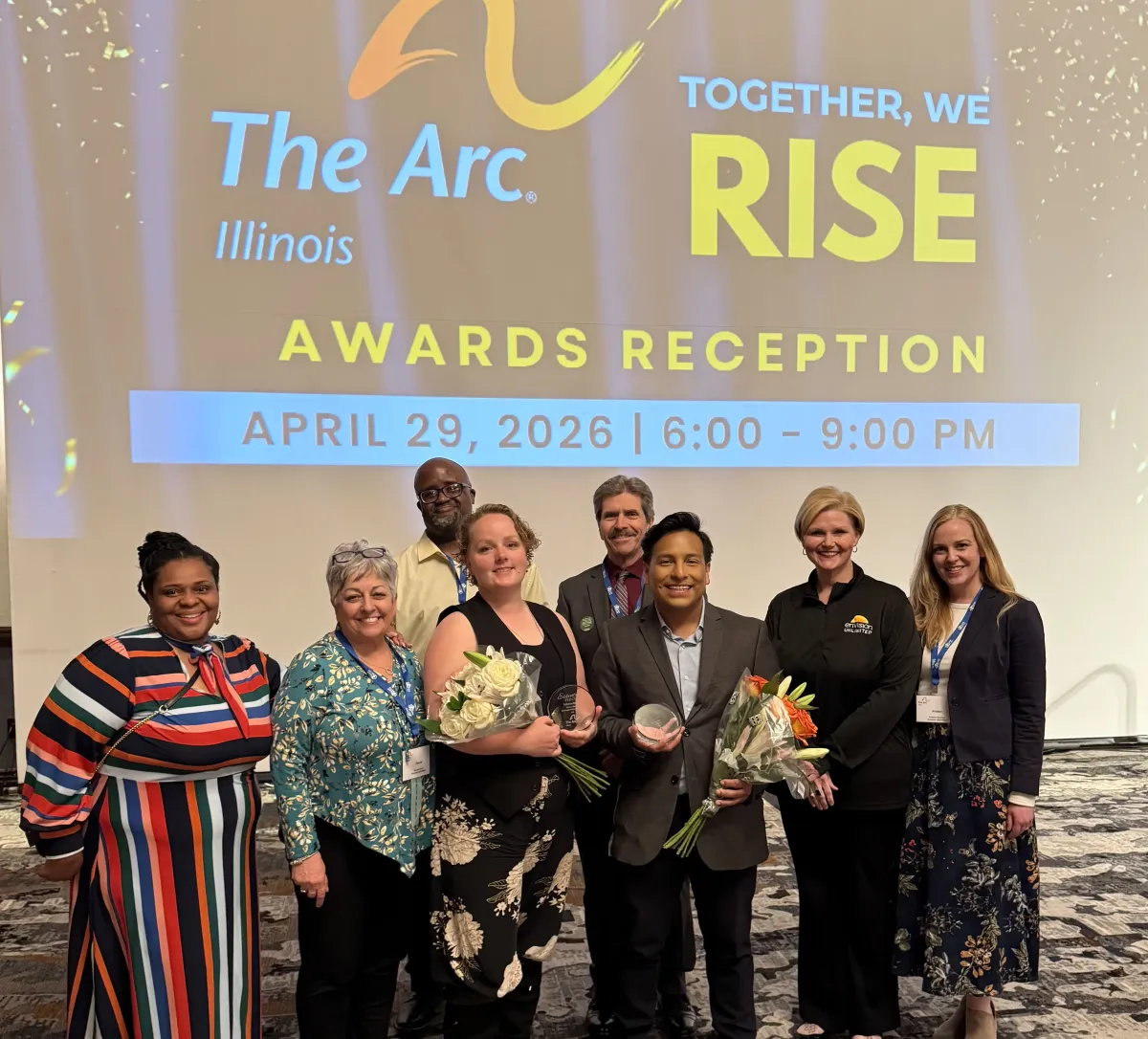 Seven professionals at The Arc Illinois 'Together, We Rise' Awards Reception pose together in front of the event banner, with two holding award flowers and glasses.