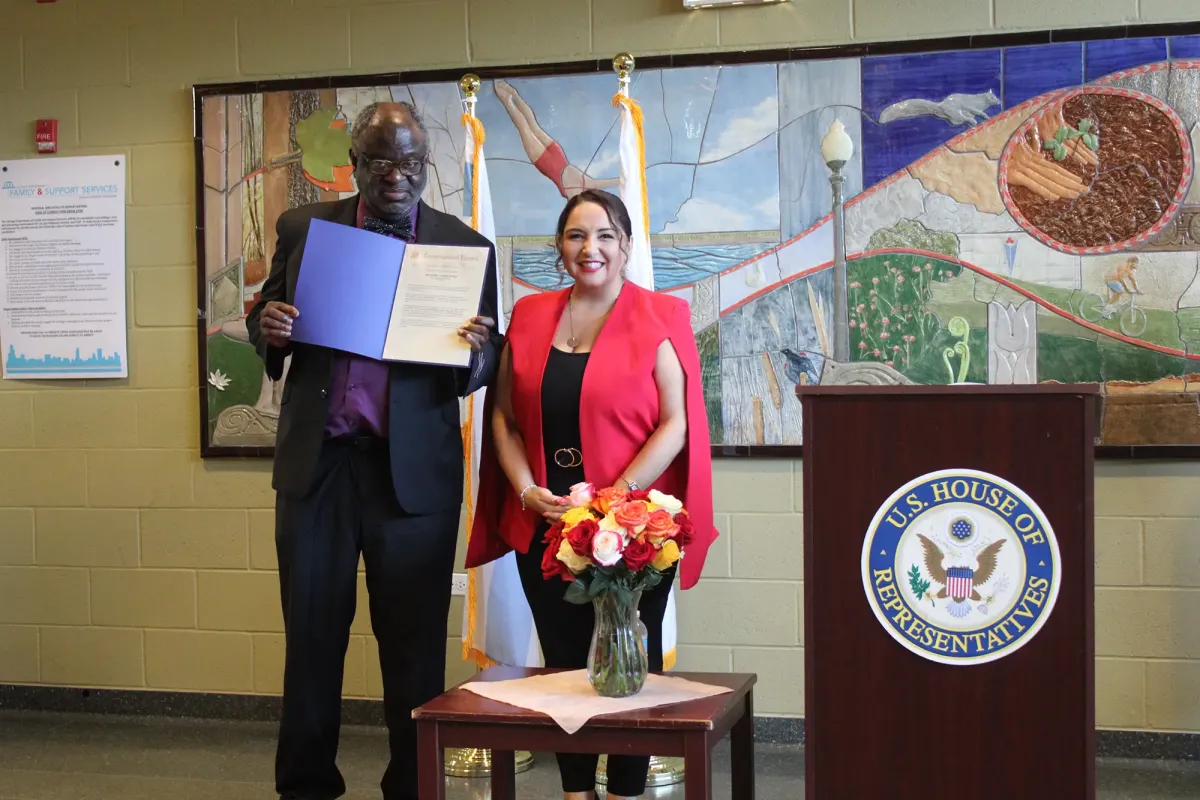 Two people standing with a U.S. House of Representatives podium and mural in the background. The man holds an open document. A vase of flowers is on a table between them.