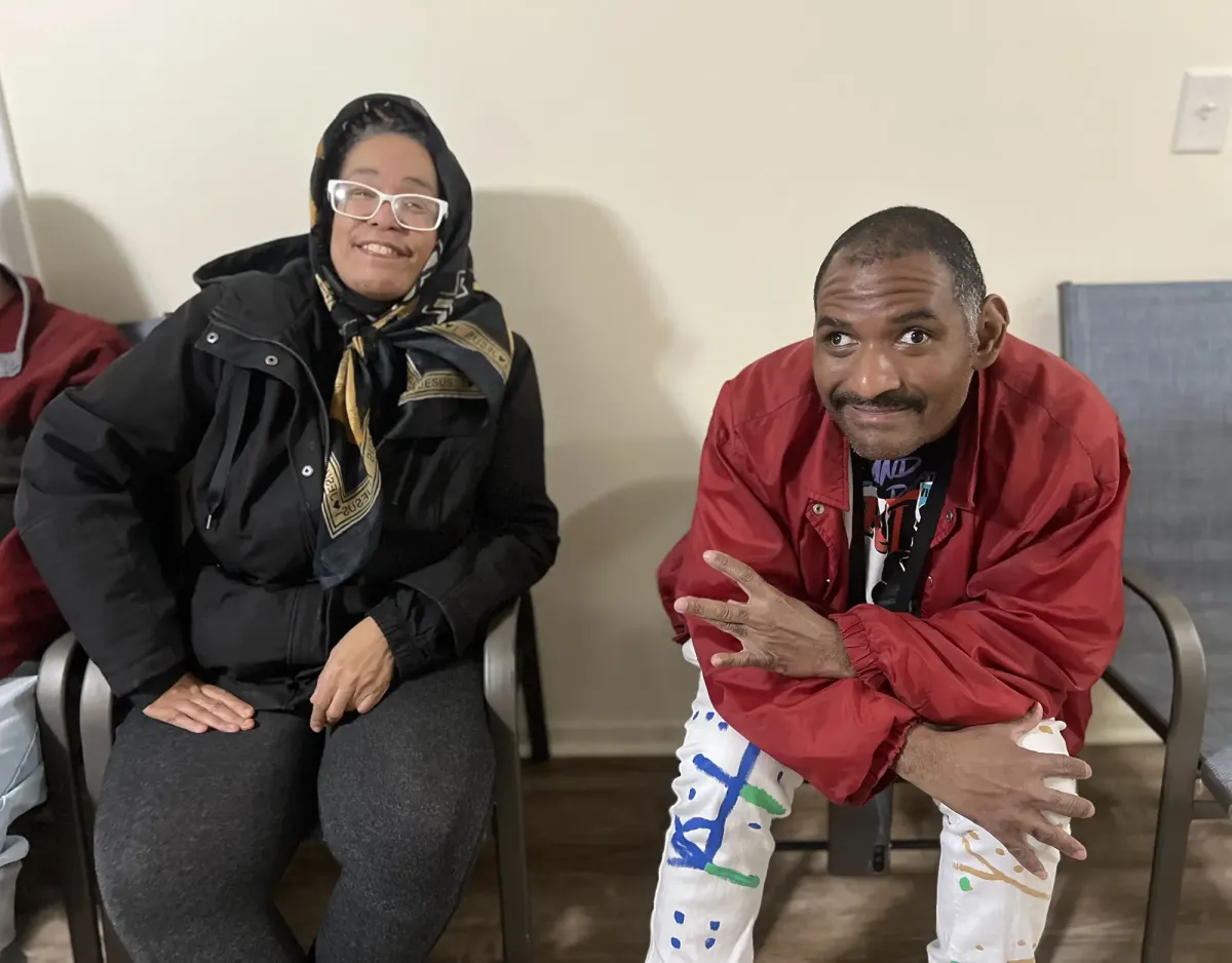Two people seated indoors, one in a headscarf and black jacket, the other in a red jacket with colorful pants.