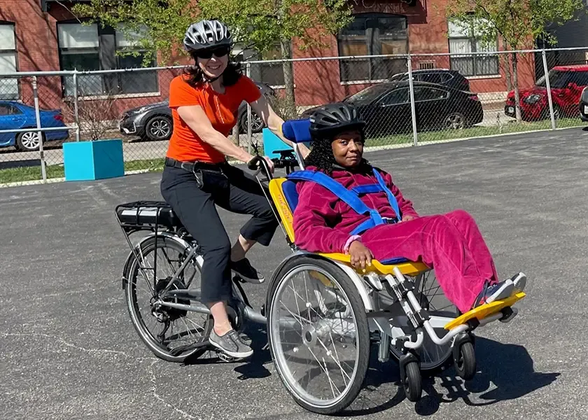 A person rides a bicycle with an attached wheelchair holding another person on a sunny day.