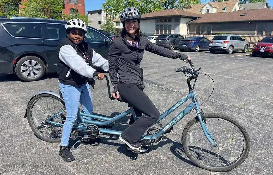 Two people riding a blue tandem bicycle in a parking lot.