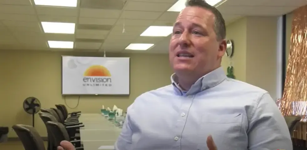 Man speaking in a conference room with a screen displaying 'Envision Unlimited.'