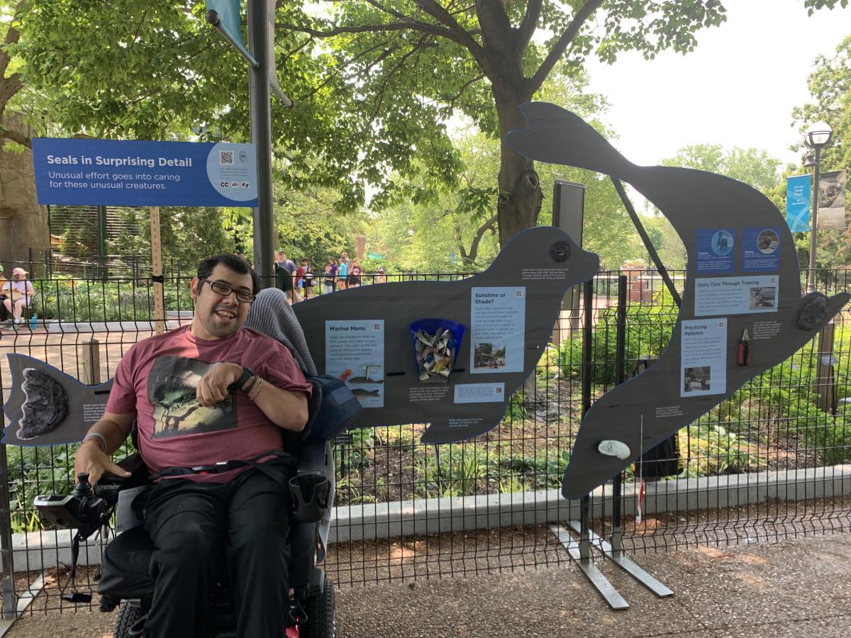 A person in a wheelchair smiles next to a seal-shaped informational display in a park.