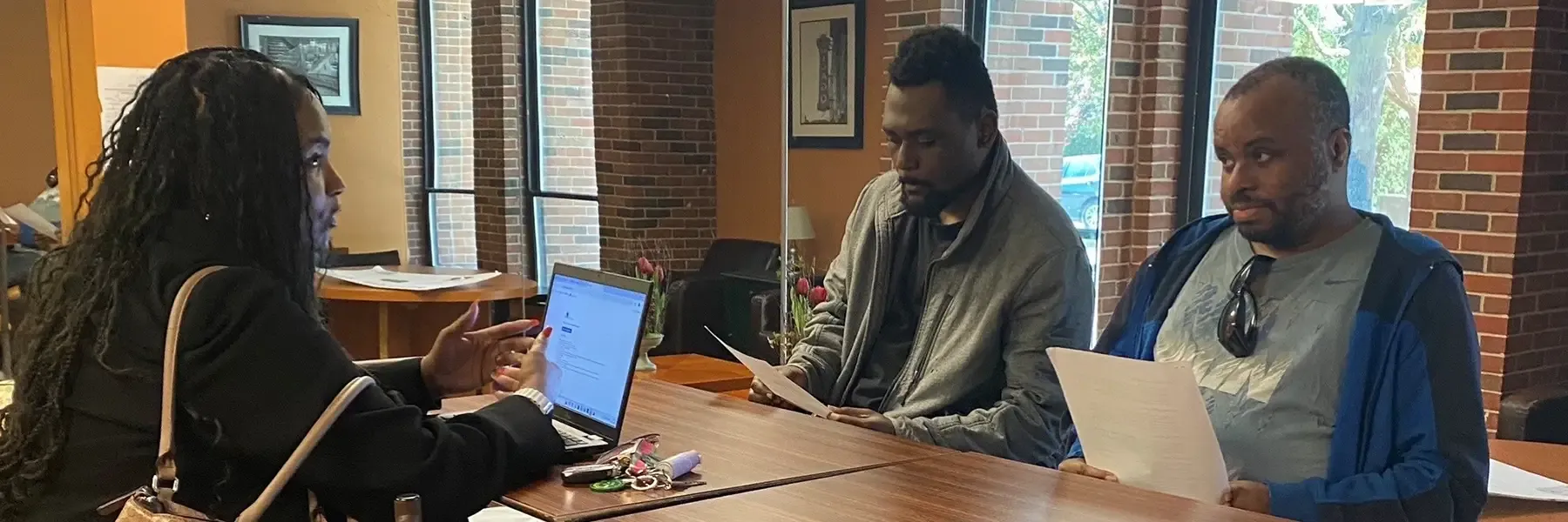 A woman with long black hair is on the left, seated at a wooden table, gesturing towards two men sitting across from her. She has a laptop open and a beige handbag placed on the back of the chair. The setting is a cozy indoor area with brick walls, large windows, and some wall art. The men are holding papers and engaging attentively.