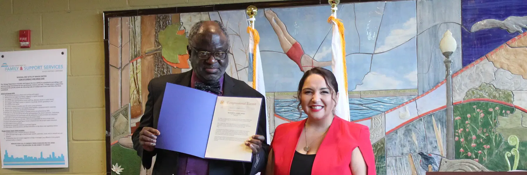 Two people standing with a U.S. House of Representatives podium and mural in the background. The man holds an open document. A vase of flowers is on a table between them.