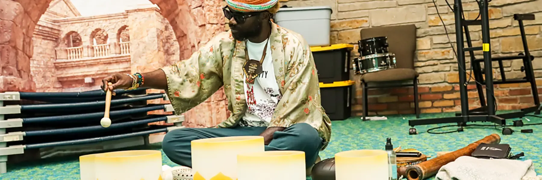 Person wearing a straw hat and patterned shirt demonstrating hand drums at a community healing event, with several yellow and white frame drums arranged on a teal and green carpeted floor.