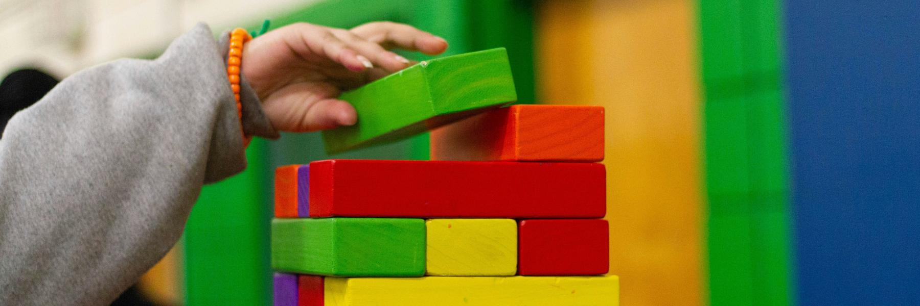 A hand wearing an orange bracelet stacks a green block onto a pile of colorful blocks.