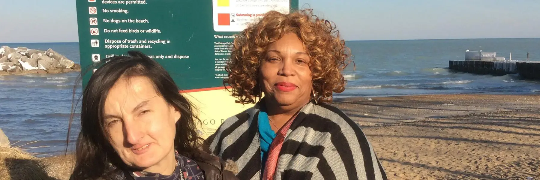Two women standing by a beach with a green informational sign behind them.