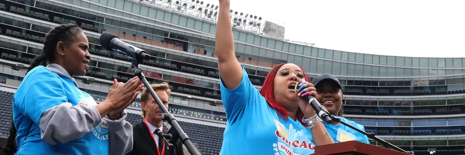 A person speaking energetically at a podium in a sports stadium with others nearby.