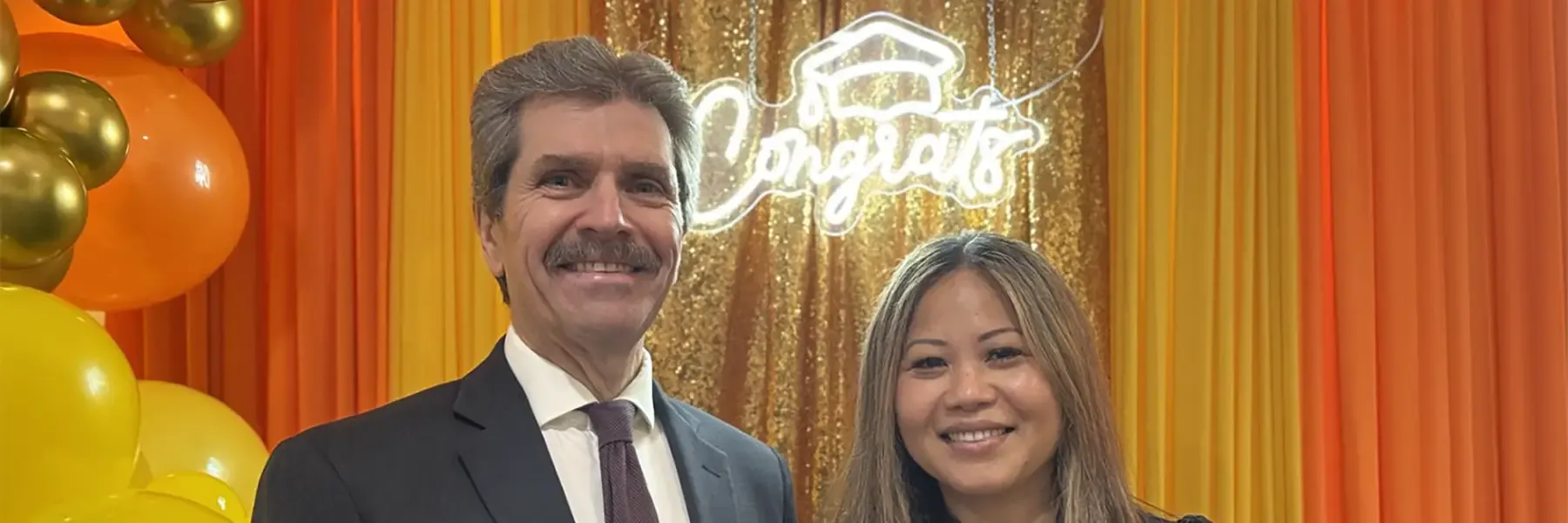 Two people smiling together at a celebratory event, posed in front of a gold sequin curtain and neon 'Congrats' sign, with colorful balloons nearby.