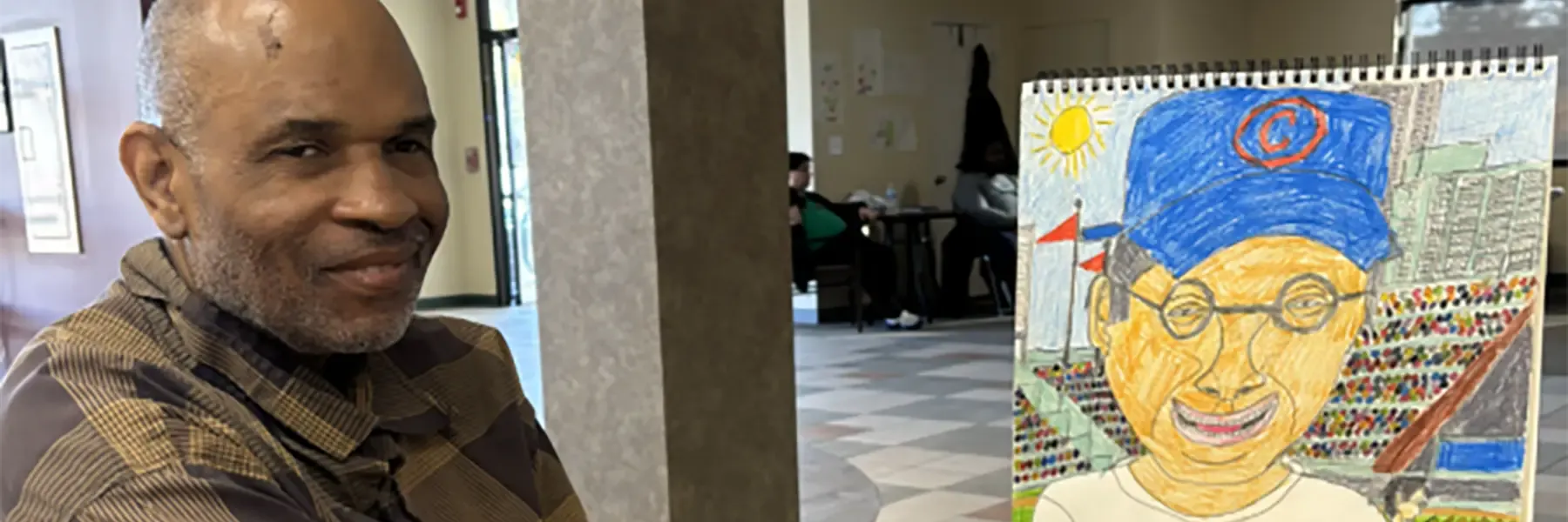 A man in a brown plaid shirt smiles while holding a colorful drawing of a person in a stadium, with colored pencils on the table.