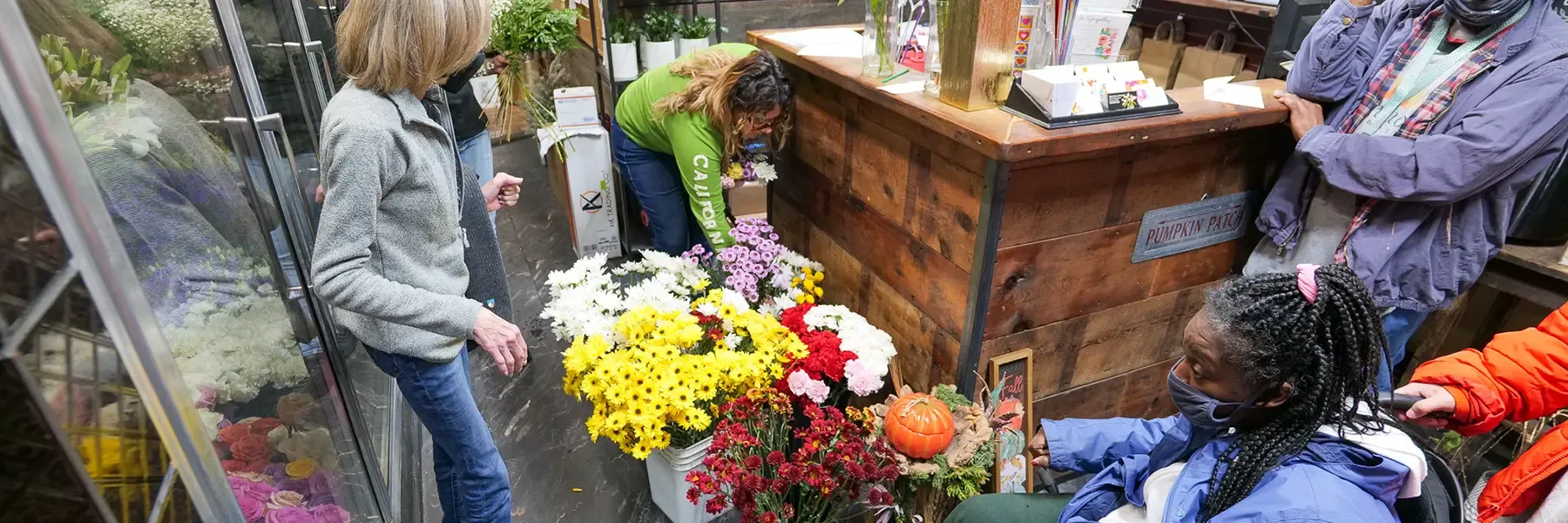 Group of people including a woman in a wheelchair admiring colorful chrysanthemums and fall flowers at a neighborhood florist shop during a community visit
