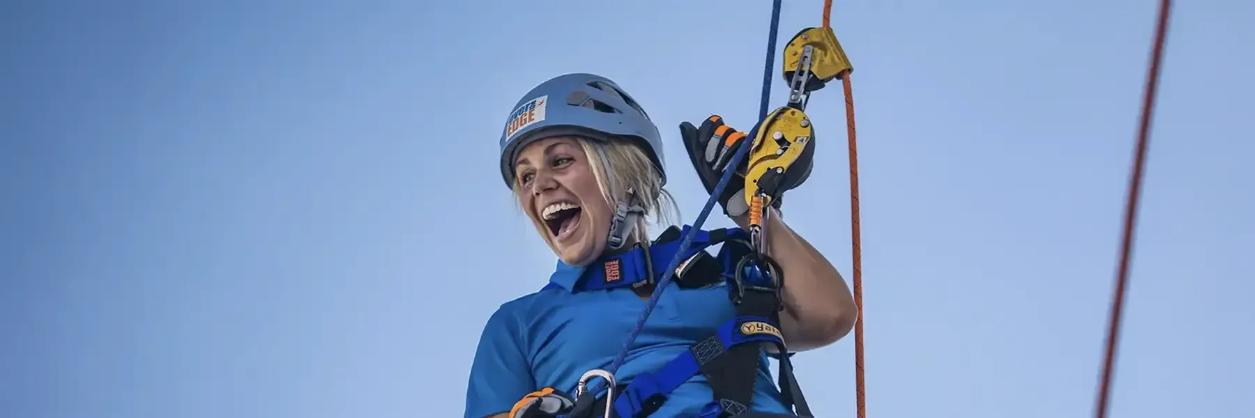 Person in blue safety gear and helmet smiling while rappelling outdoors, holding yellow and orange climbing carabiners attached to secured rope lines.