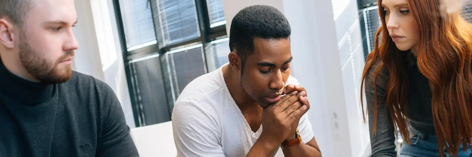 Three people in a quiet, reflective moment together in a bright office space, with the center figure appearing thoughtful while others sit attentively nearby.