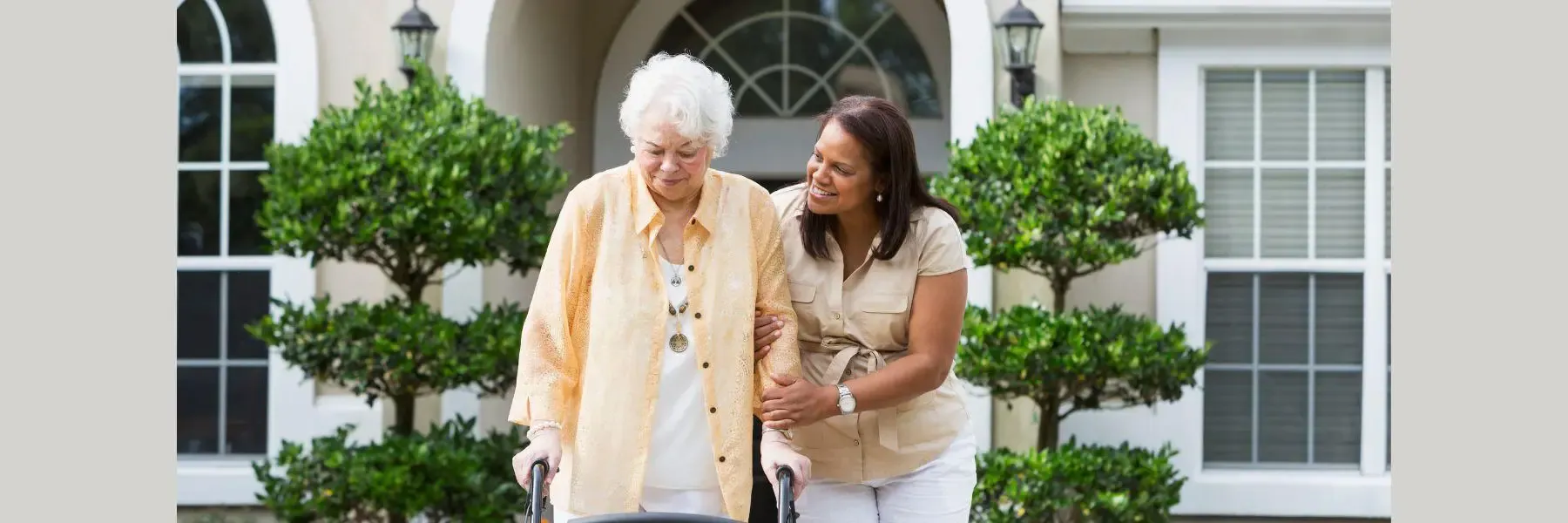 Older woman using a walker walks arm-in-arm with a smiling caregiver outside an elegant residential home