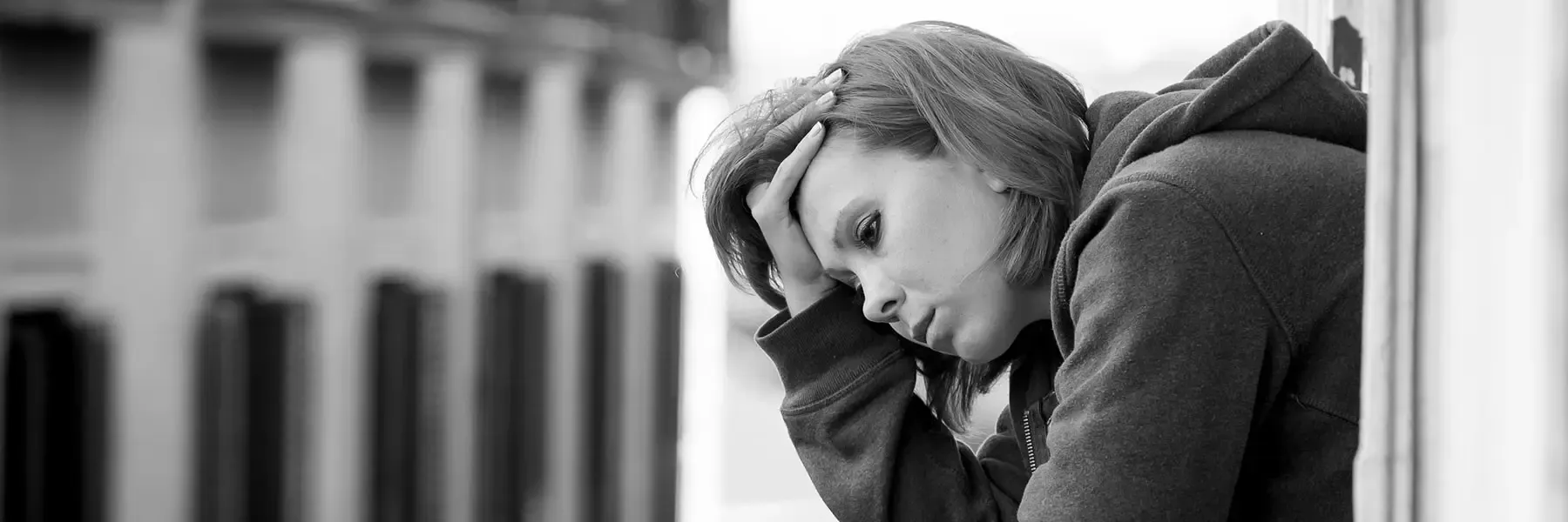 The black and white image shows a person leaning against a wall, looking down with a somber expression. They are wearing a hooded sweatshirt, with the hood down. Their hand is resting on the side of their head, fingers threaded through their hair. The background is blurred, suggesting an urban setting with tall buildings. The grayscale tones emphasize the solemn mood of the scene.