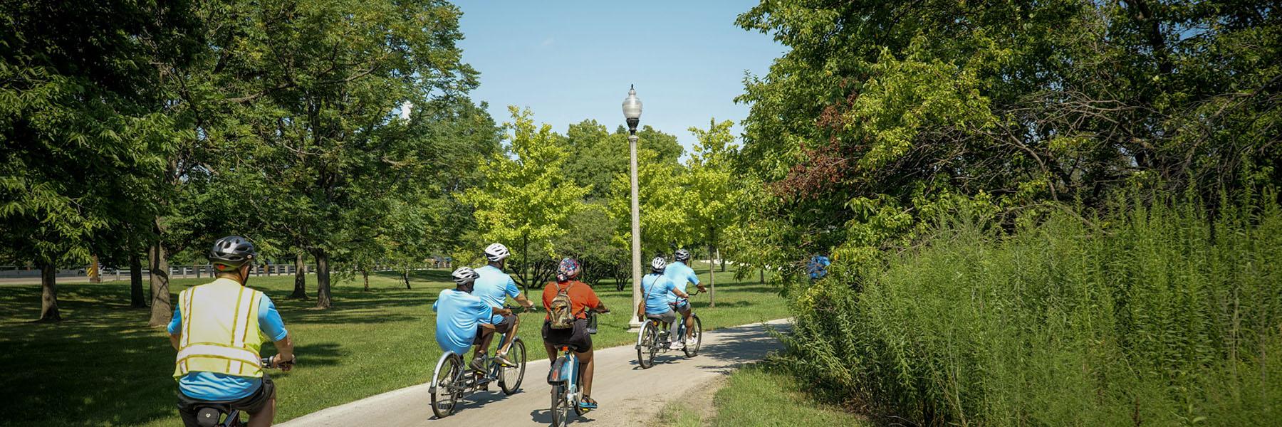 A group of cyclists bikes along a lakefront trail surrounded by green foliage and nature