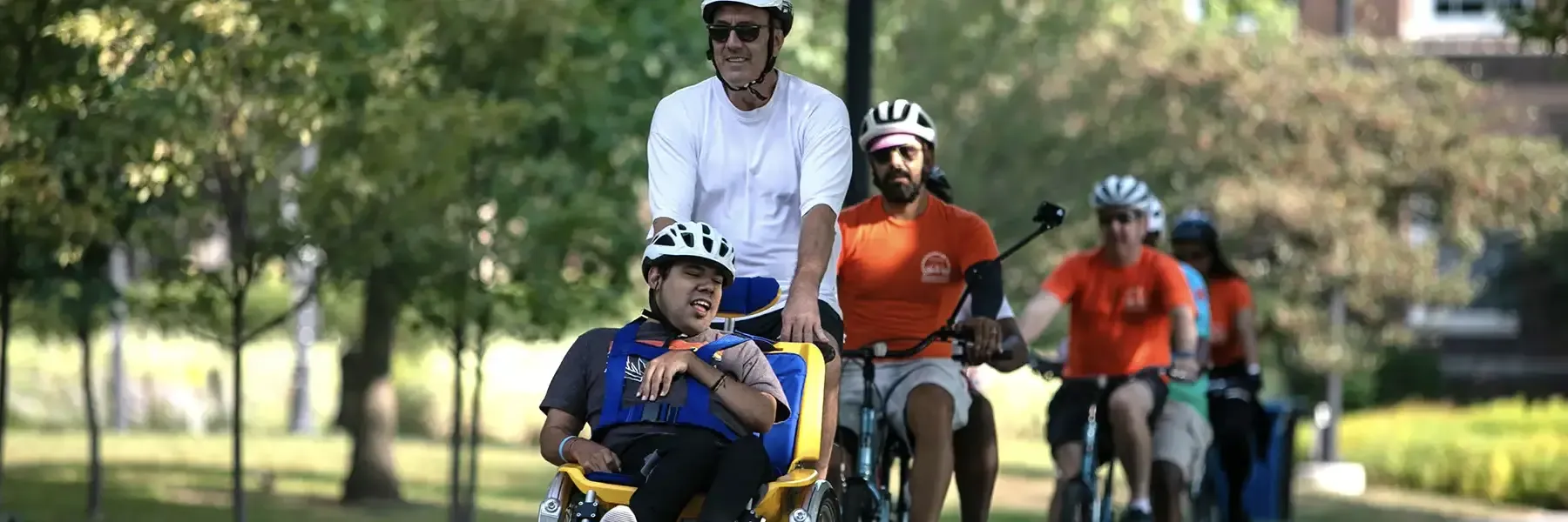 An adult cyclist leads a group ride while towing a child in a yellow cargo bike trailer on a sunny park path. Multiple cyclists in orange jerseys follow behind on the tree-lined, paved route.