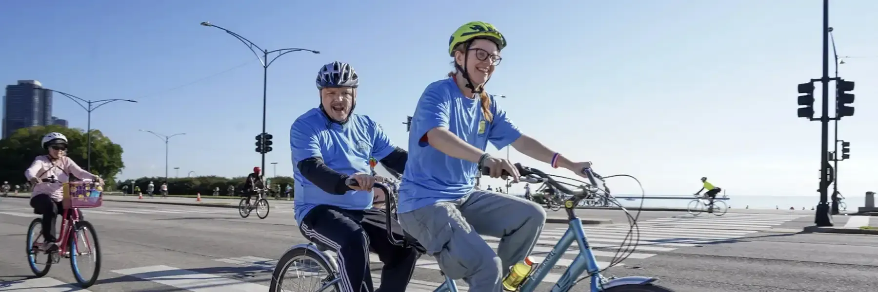 Two cyclists in blue shirts riding a blue tandem bicycle on a city street with downtown high-rise buildings in the background, traffic signals, and other cyclists visible.