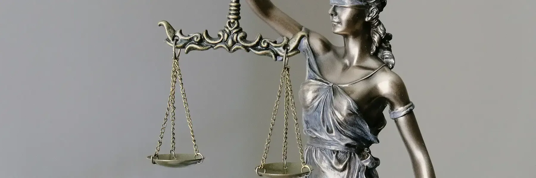 Metallic statue of Lady Justice in profile, holding balanced scales of justice in both hands, wearing a crown and classical robes against a neutral gray background.