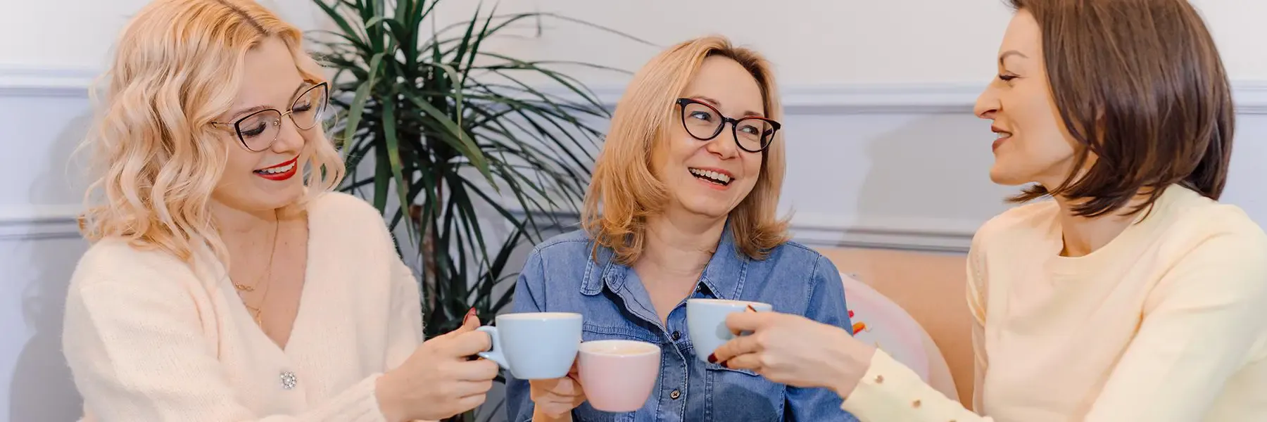 Three women smiling and toasting with coffee cups in an indoor office setting, conveying a moment of workplace camaraderie and celebration.