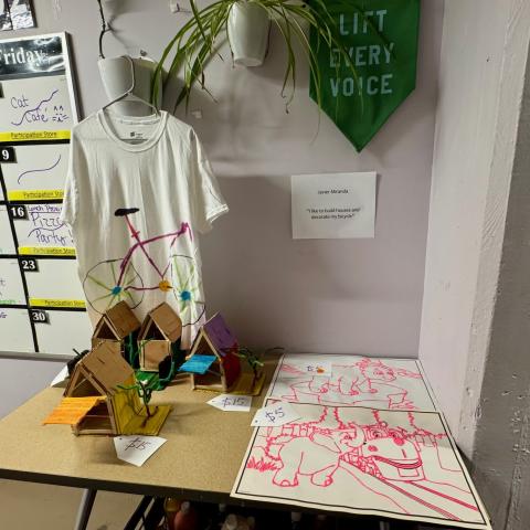 A display of artwork that includes a handpainted t-shirt, bird houses and drawings.