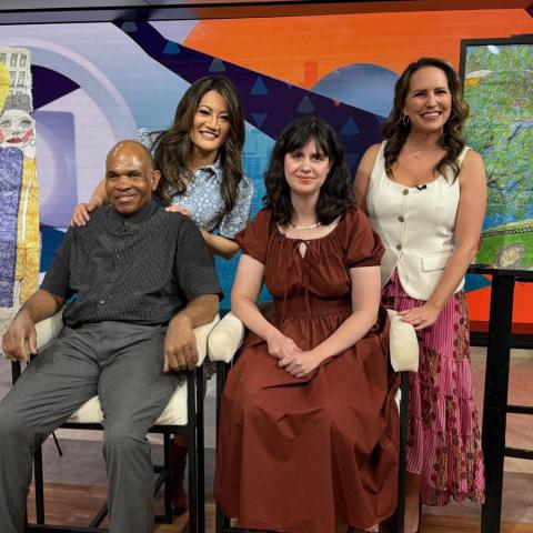 A group of four people sit for a photo at a news studio with artwork on display behind them.